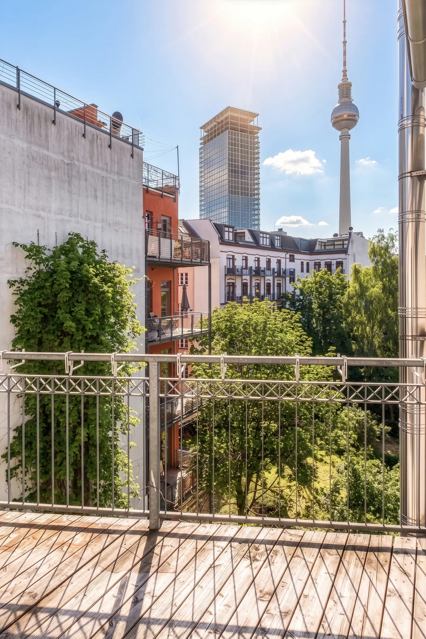 View from a wooden balcony with a metal railing overlooking Berlin buildings and the TV Tower on a sunny day.
