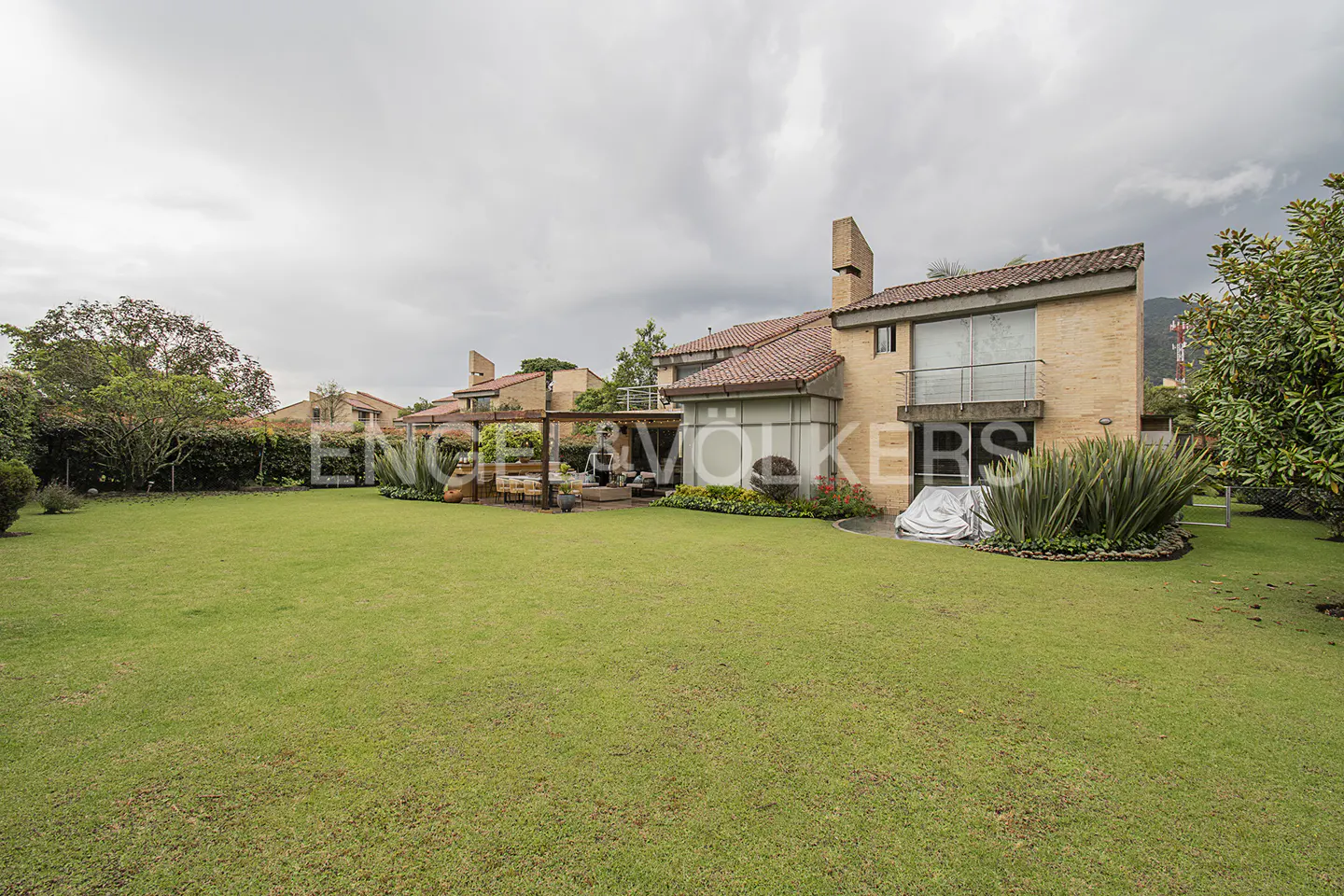 Backyard view of a two-story brick house with a red tile roof and a green lawn under a cloudy sky.