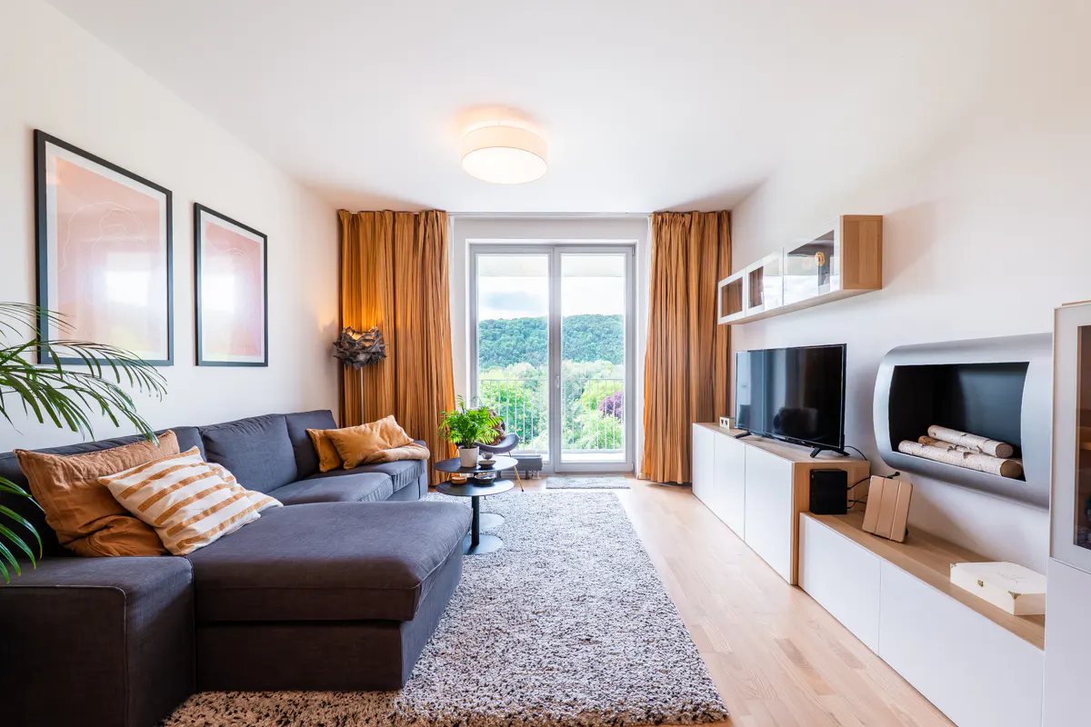 Modern living room with gray sectional sofa, brown curtains, and a view of green trees through a glass door.