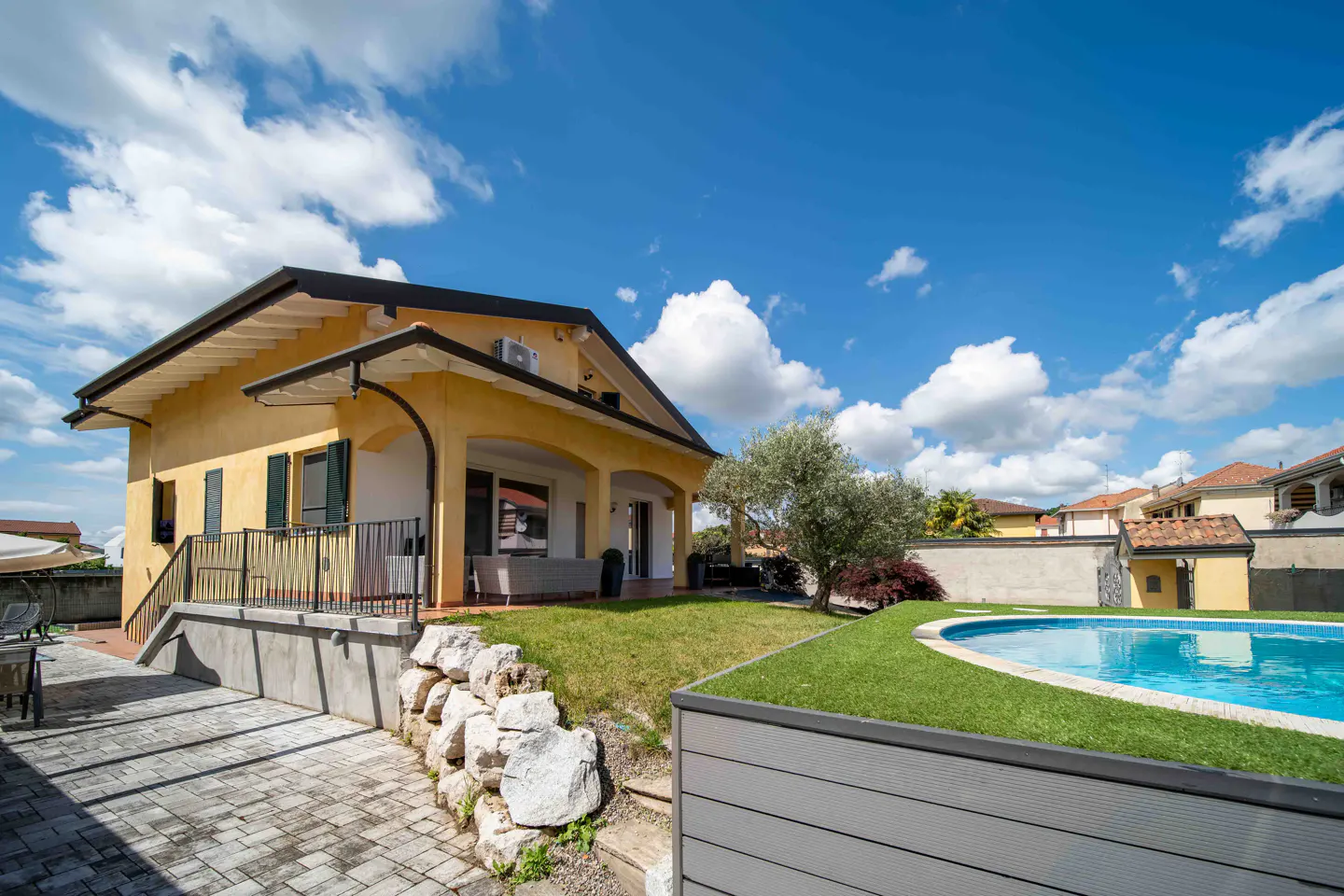 Exterior view of a yellow two-story house with a pool, lawn, and stone retaining wall under a blue sky with white clouds.