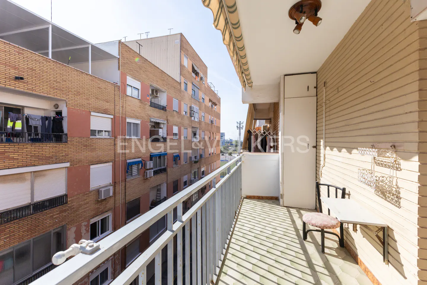 A narrow apartment balcony with a small table and stool, overlooking a neighboring brick apartment building.