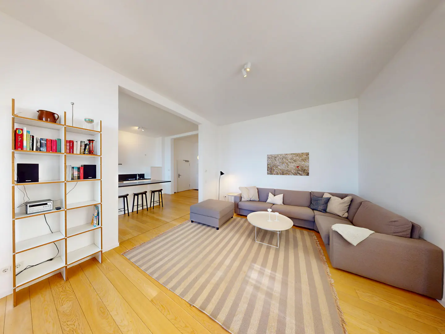 Bright living room with a gray sectional sofa, striped rug, and a bookshelf filled with books. Kitchen in the background.