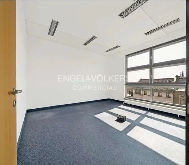Empty office space with blue carpet, white walls, and large window. Engel & Völkers Commercial logo on the wall.