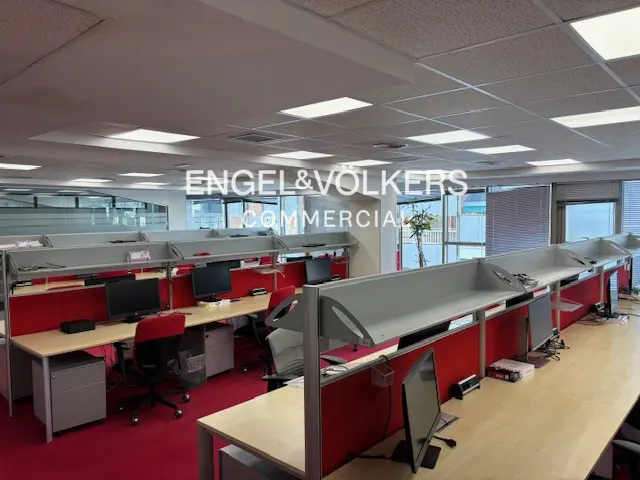 Bright office space with rows of desks, red partitions, and computer setups. Large windows offer natural light.