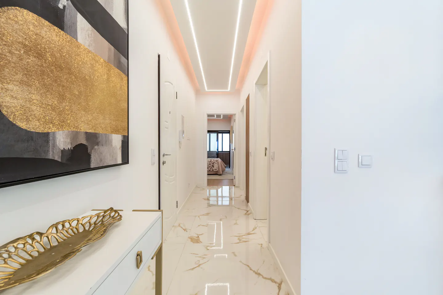 Modern hallway with marble floors, gold-accented art, a white console table, and recessed linear ceiling lighting.