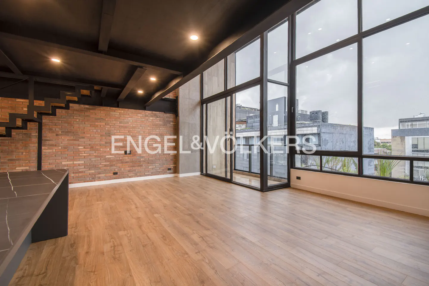Modern loft with brick wall, wood floors, black ceiling, and floor-to-ceiling windows overlooking a city skyline.