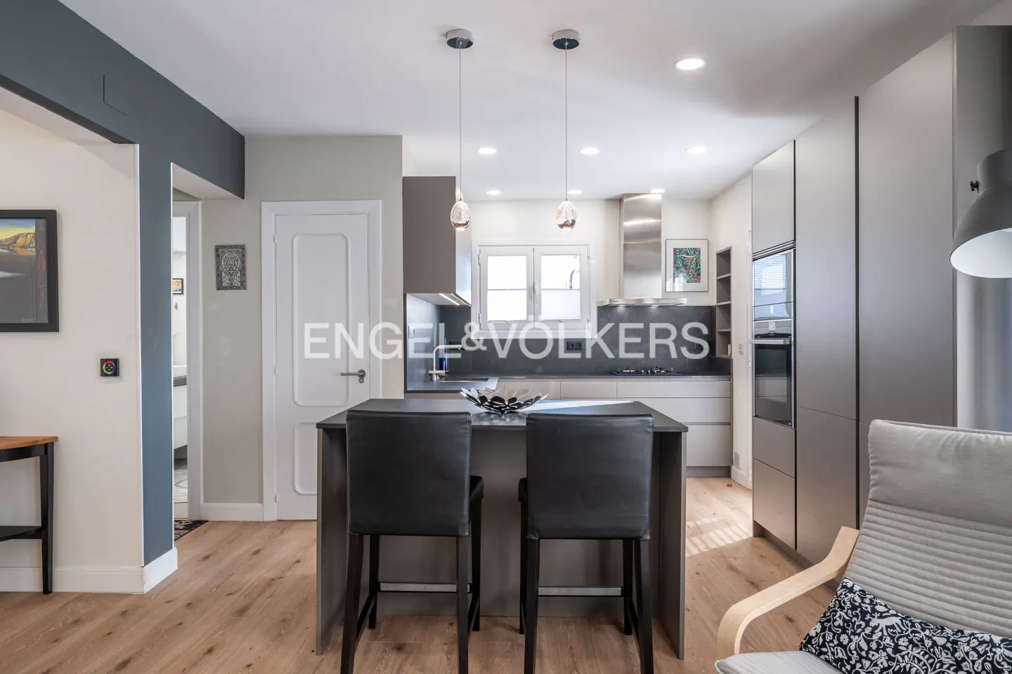 A modern kitchen with gray cabinets, a central island with two black stools, and light wood floors.