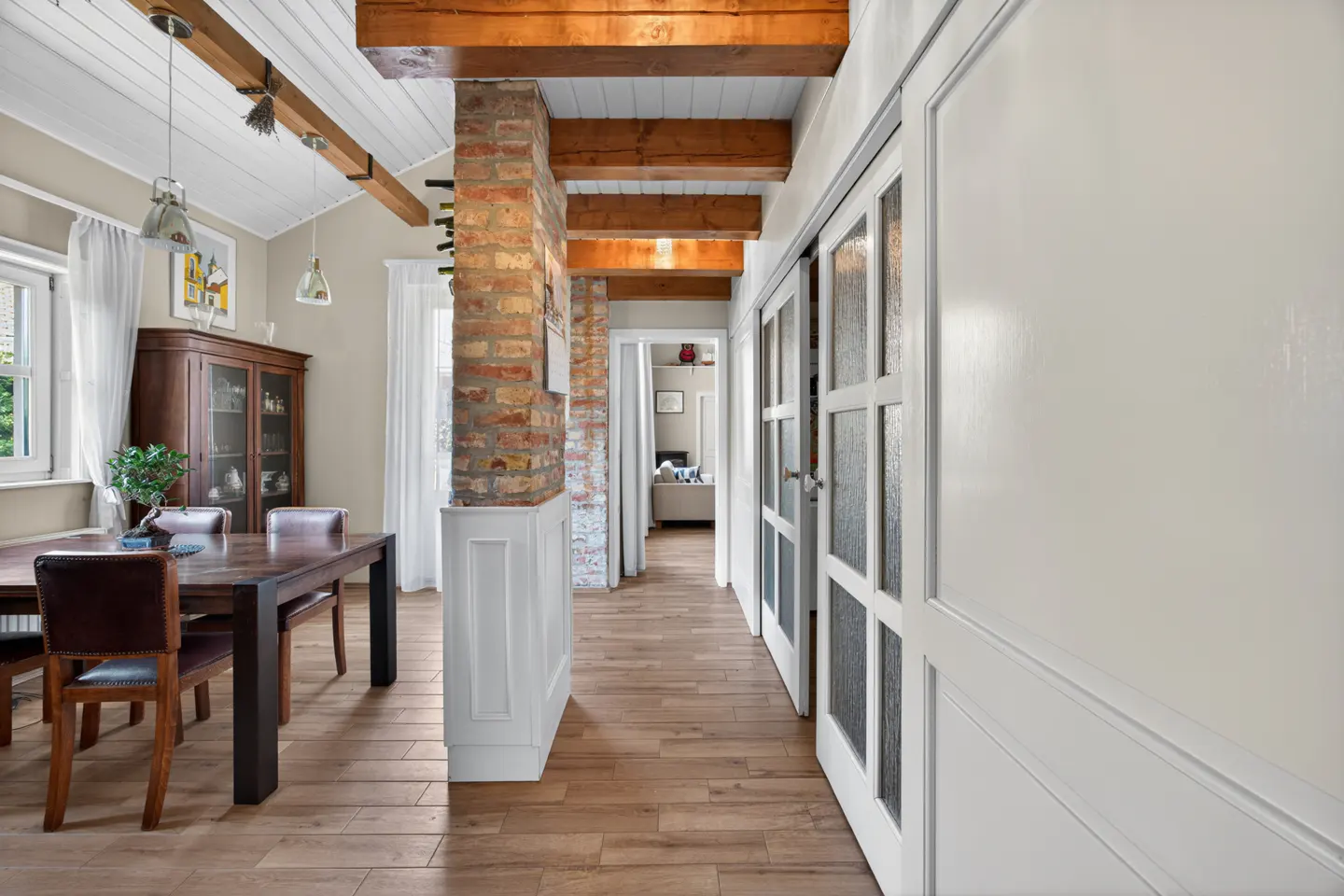 Interior view of a dining room and hallway with wood floors, exposed brick, and wooden beams. A dark wood table and chairs are on the left. White sliding doors are on the right.