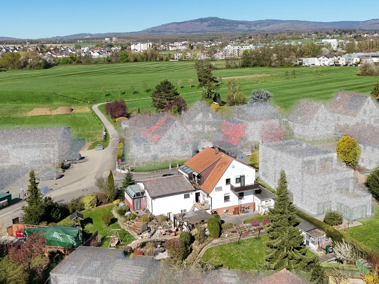 Aerial view of a white house with an orange roof, surrounded by green lawns and trees, with a field and mountains in the background.