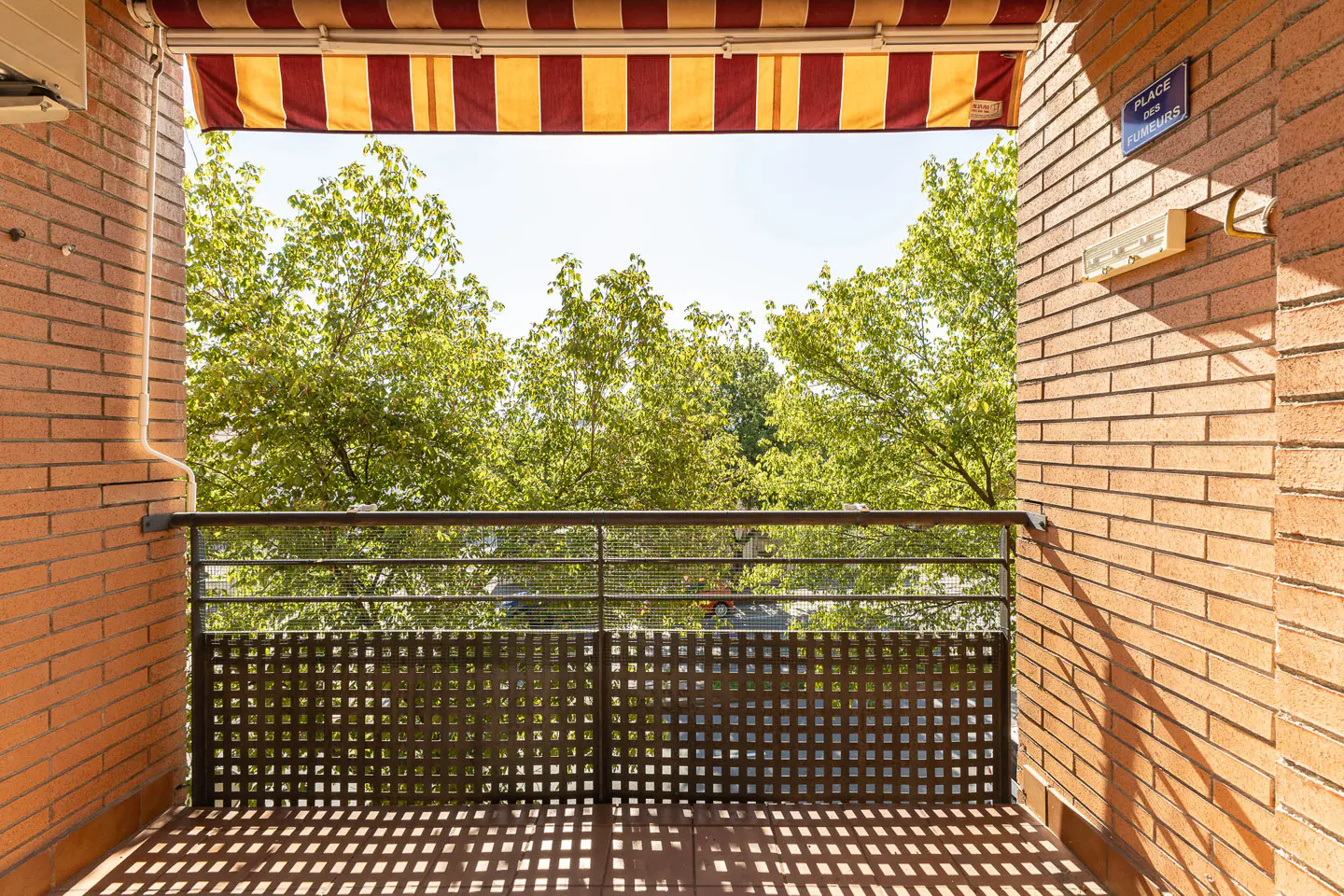 Balcony view with brick walls, metal railing, and a red and yellow striped awning. Green trees are visible in the background.