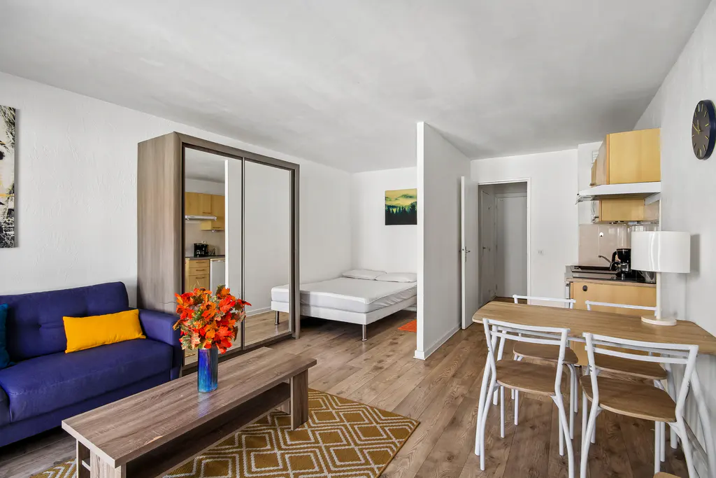 Bright studio apartment with wood floors, a blue sofa, a bed, a dining table, and a small kitchen.