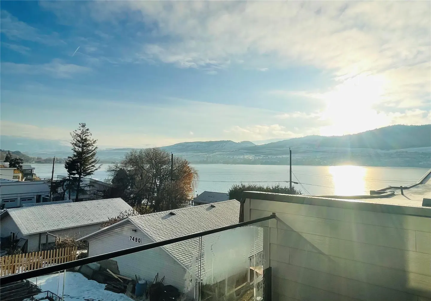 A winter view from a balcony shows snow-covered rooftops, a lake reflecting sunlight, and distant mountains under a blue sky.