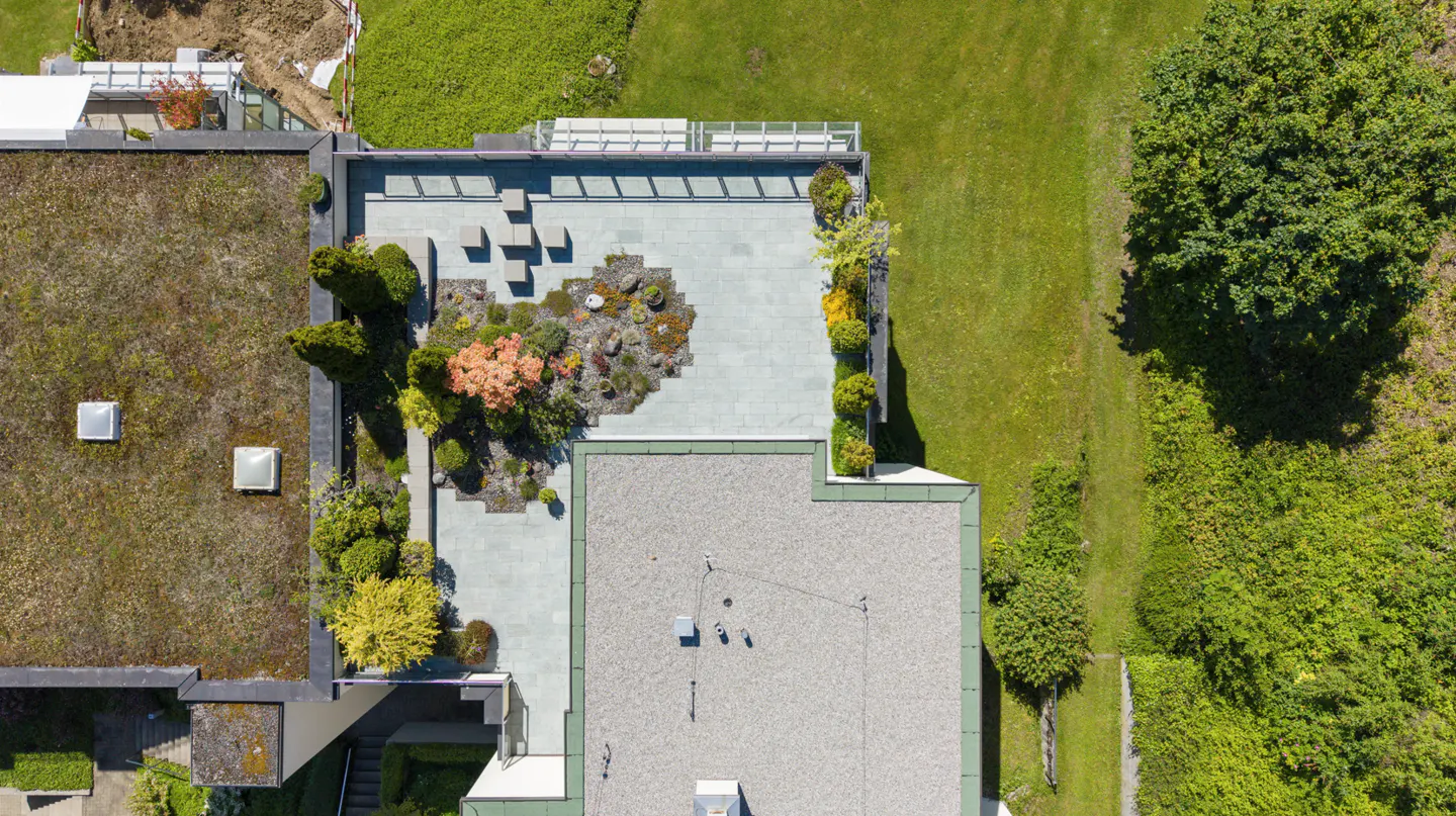 Aerial view of a modern home with a green roof, a stone patio with small trees, and a gravel roof next to a grassy lawn.