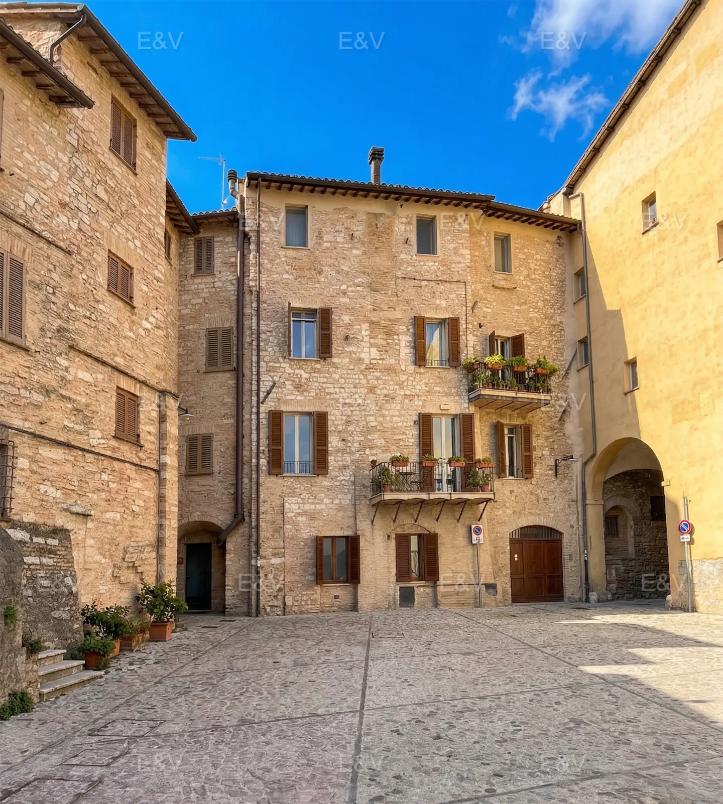 Stone buildings surround a cobblestone square under a blue sky. Balconies with flowers adorn the buildings. Brown shutters accent the windows.