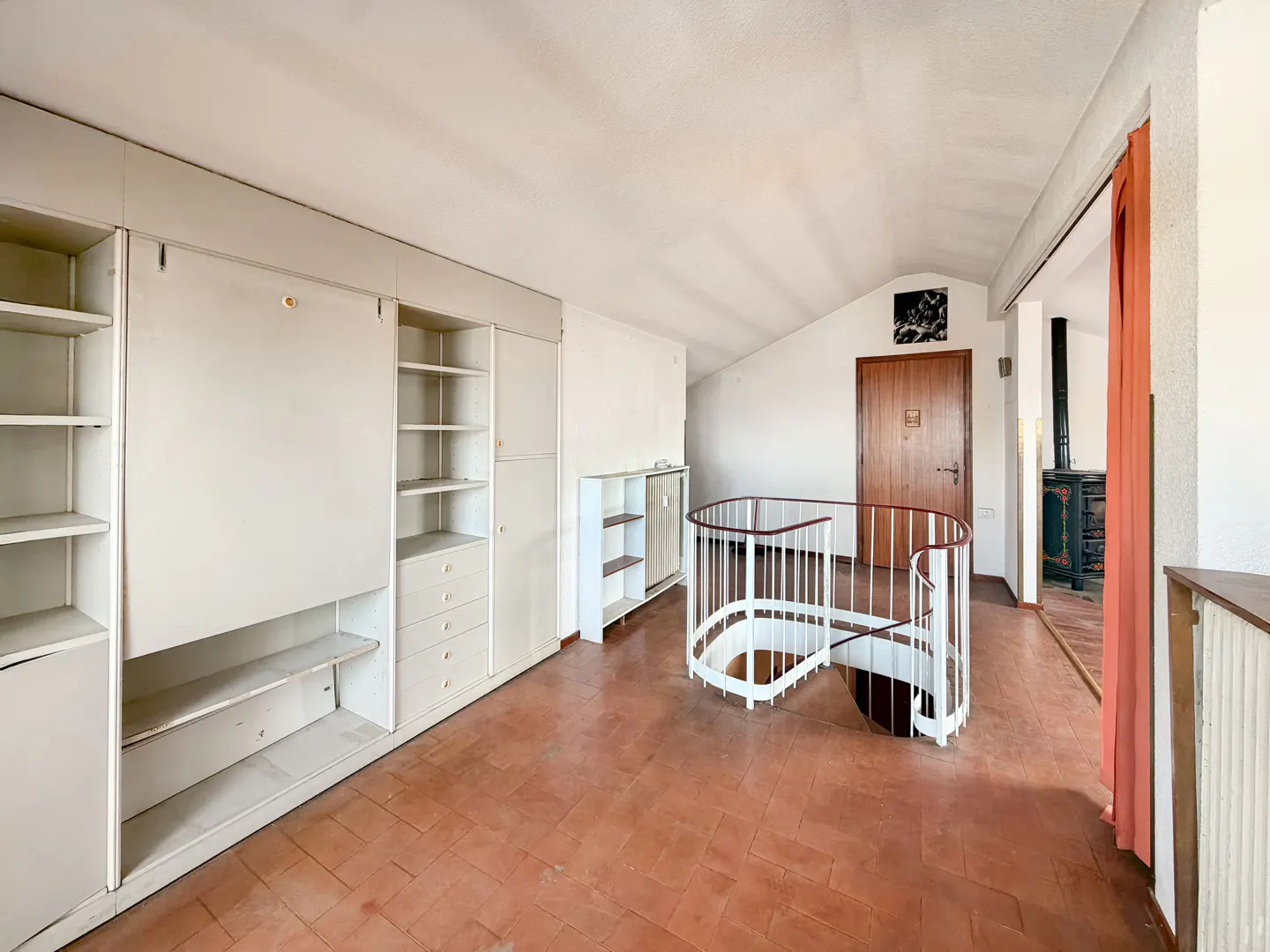 Attic room with terracotta tile floor, white built-in shelving, and a white spiral staircase with brown handrail.