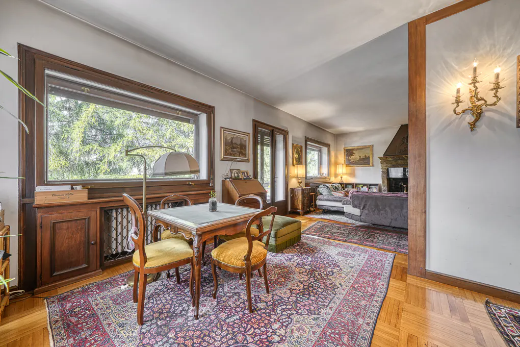 A living room with a table, chairs, and a large window overlooking trees. The room has a fireplace and a patterned rug.