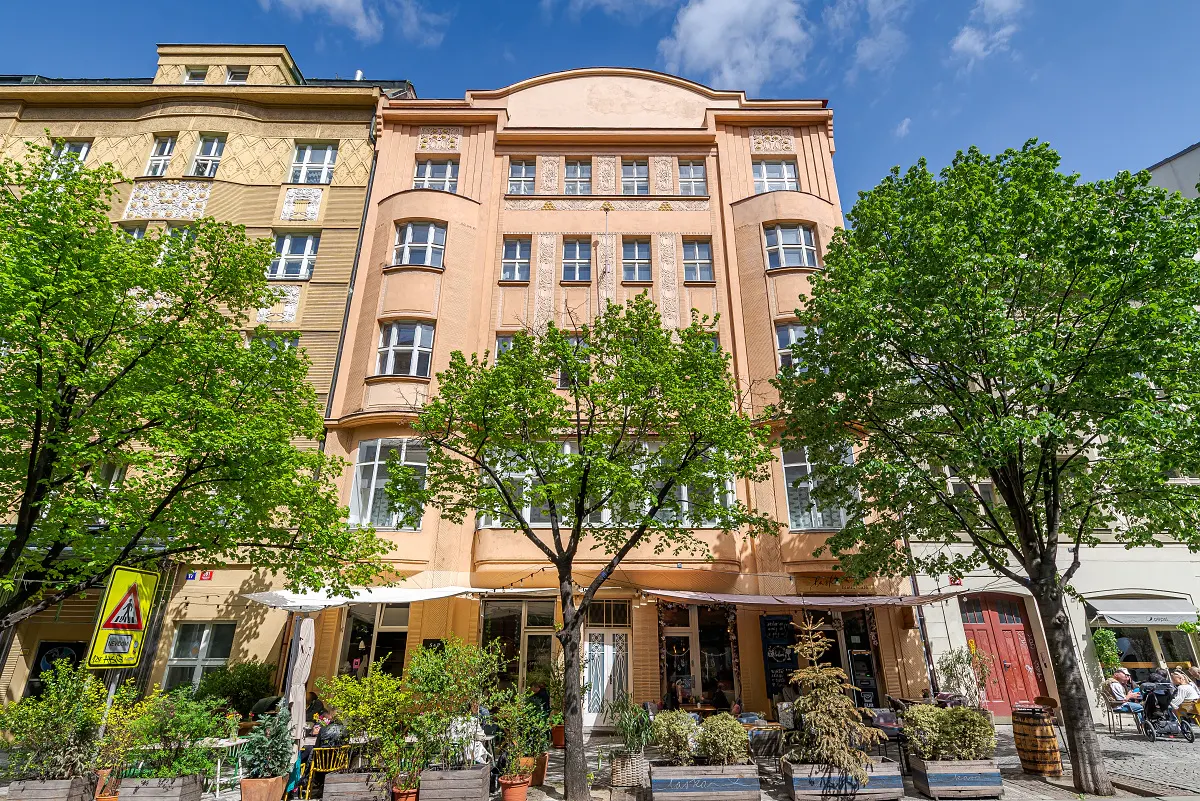 Exterior view of a peach-colored, multi-story building with white-framed windows, trees, and outdoor seating.