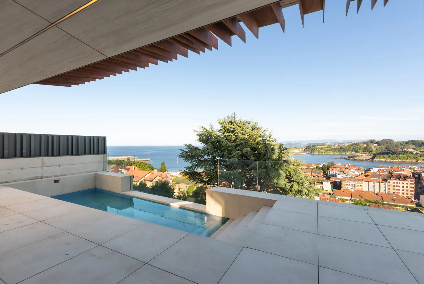 Modern terrace with a small rectangular pool overlooking a coastal town, blue ocean, and a large tree under a clear sky.
