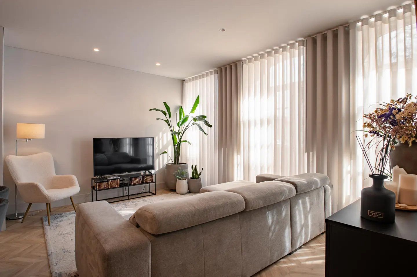 Bright living room with beige sofa, white armchair, TV, plants, and sheer curtains.