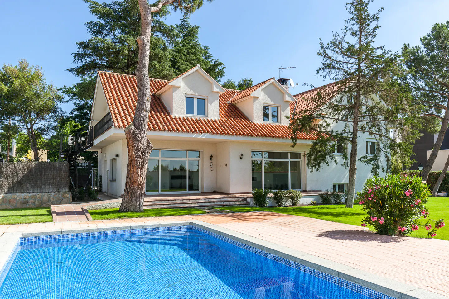 White two-story house with a red tile roof, large windows, and a blue swimming pool in a sunny backyard with trees.