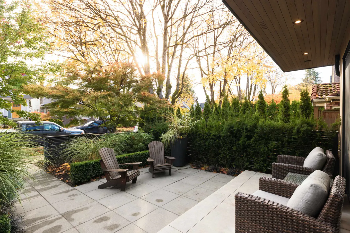 Outdoor patio with two Adirondack chairs, wicker furniture, and green hedges. Sunlight filters through trees in the background.