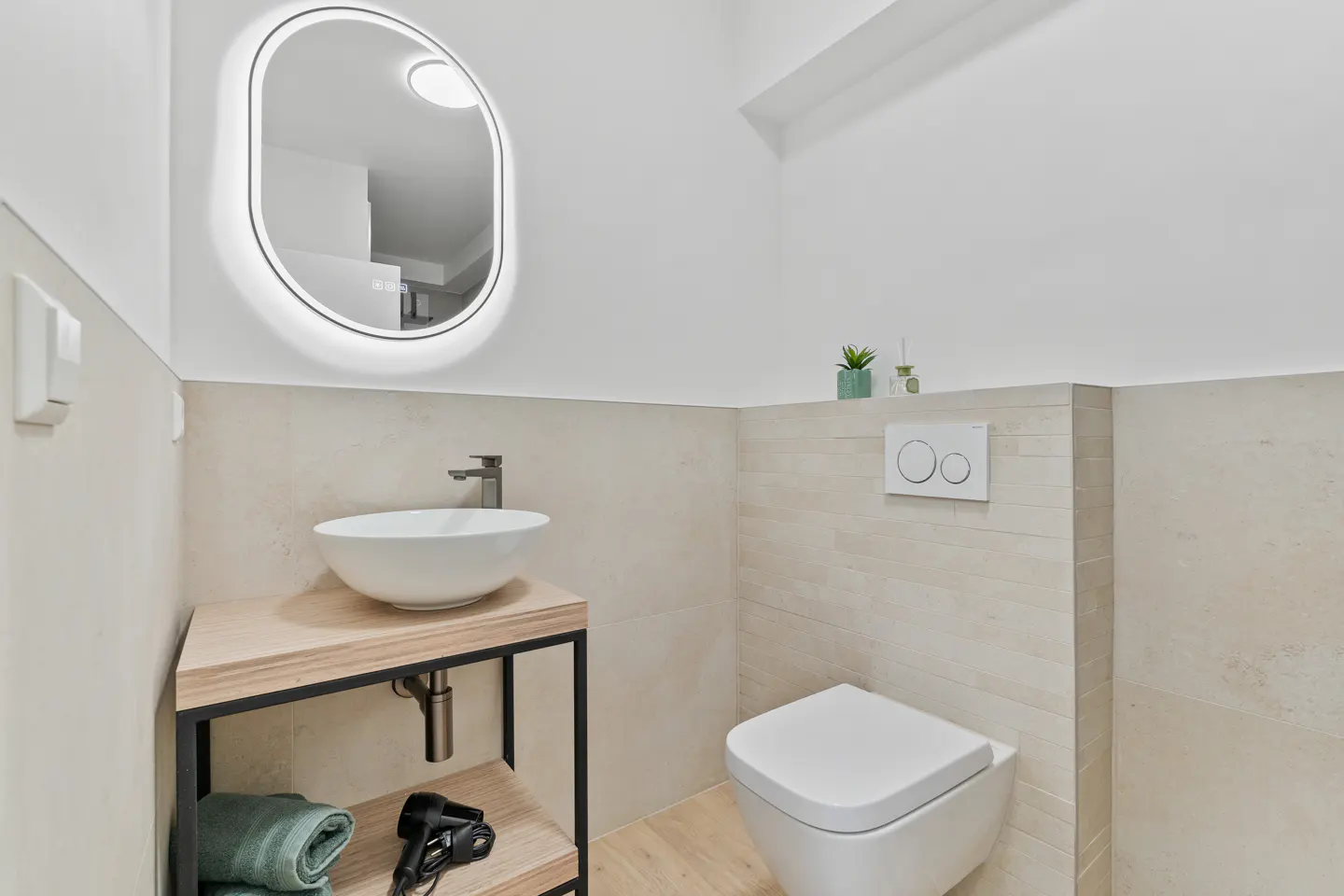 Bright bathroom with white walls, beige tile, and wood floors. A white sink sits on a wood and black metal stand, next to a white toilet. A lighted mirror hangs above the sink.