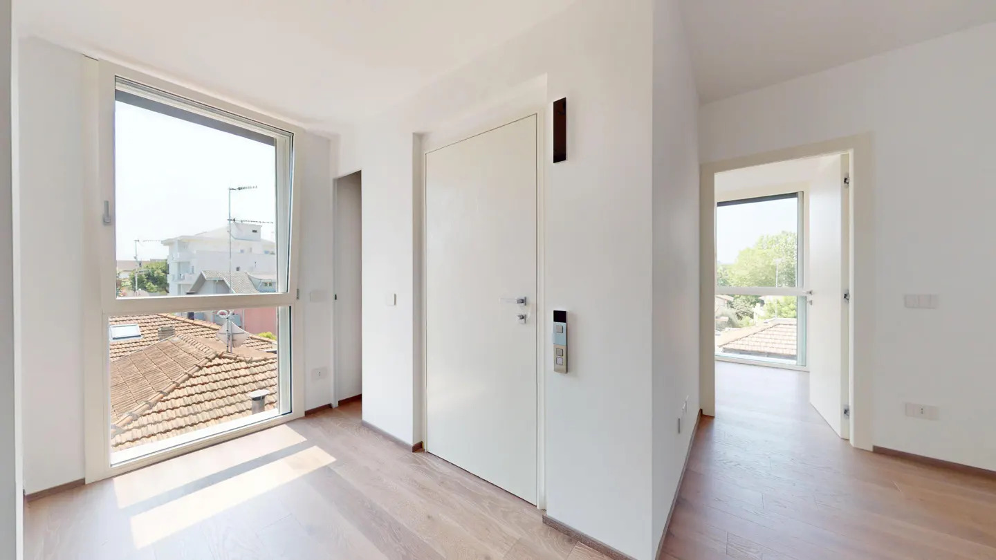 Bright, modern hallway with light wood floors, white walls, and two large windows showing rooftops and trees. A white door is centered.