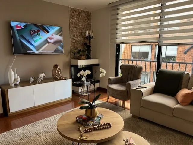 Modern living room with a beige sofa, armchair, round wooden coffee table, TV, and a fireplace with stone accents.