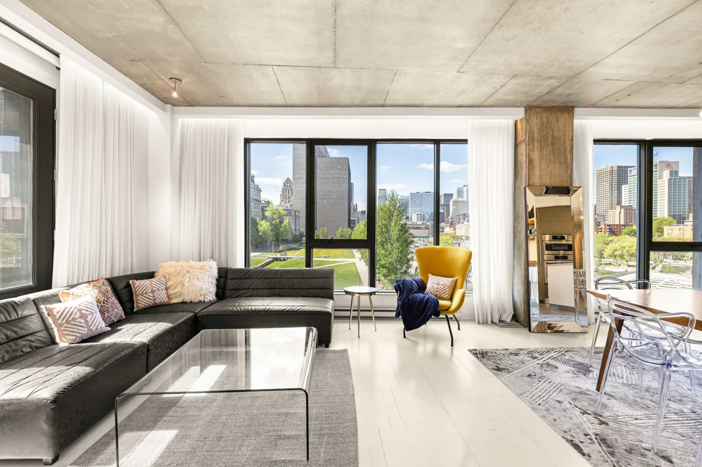 Bright, modern living room with a black sectional, yellow chair, and city views from large windows. Concrete ceiling and white floors.