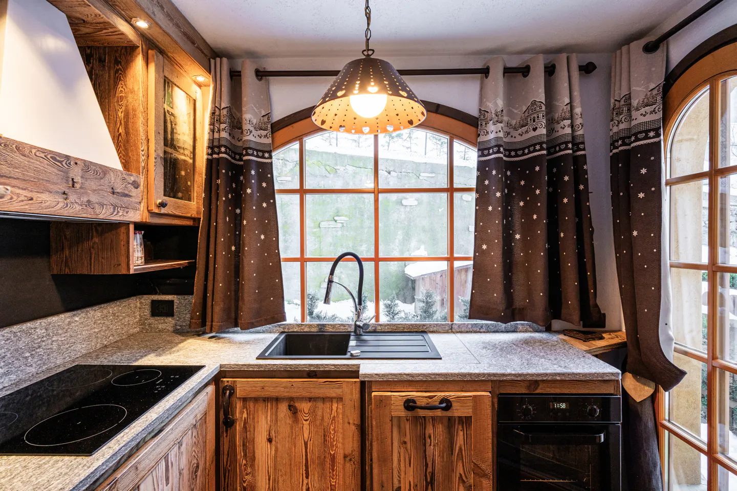 Rustic kitchen with wood cabinets, granite counters, and a black sink under a window with patterned curtains.