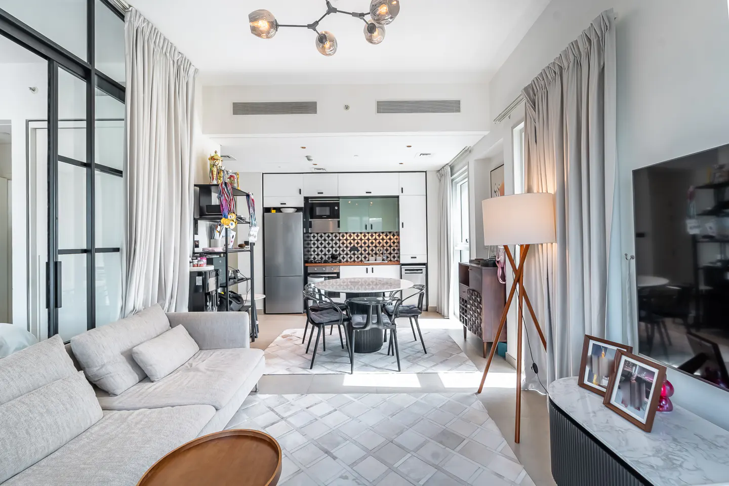Modern open-plan living room with a grey sofa, dining area, and kitchen featuring white cabinets and patterned backsplash.