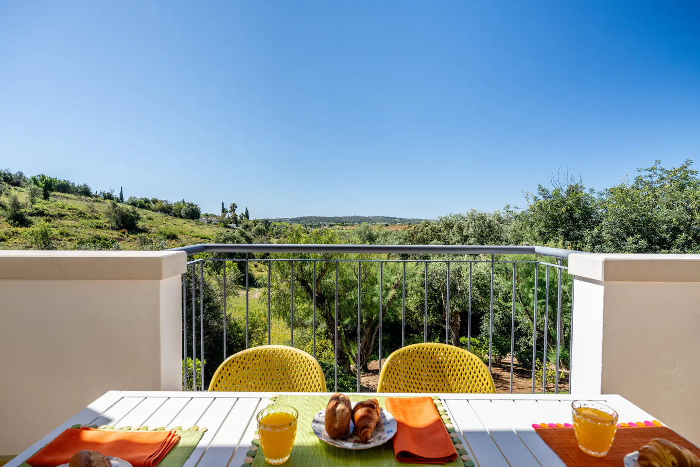 Balcony view with breakfast. White table, yellow chairs, juice, croissants. Green hills and blue sky in the background.