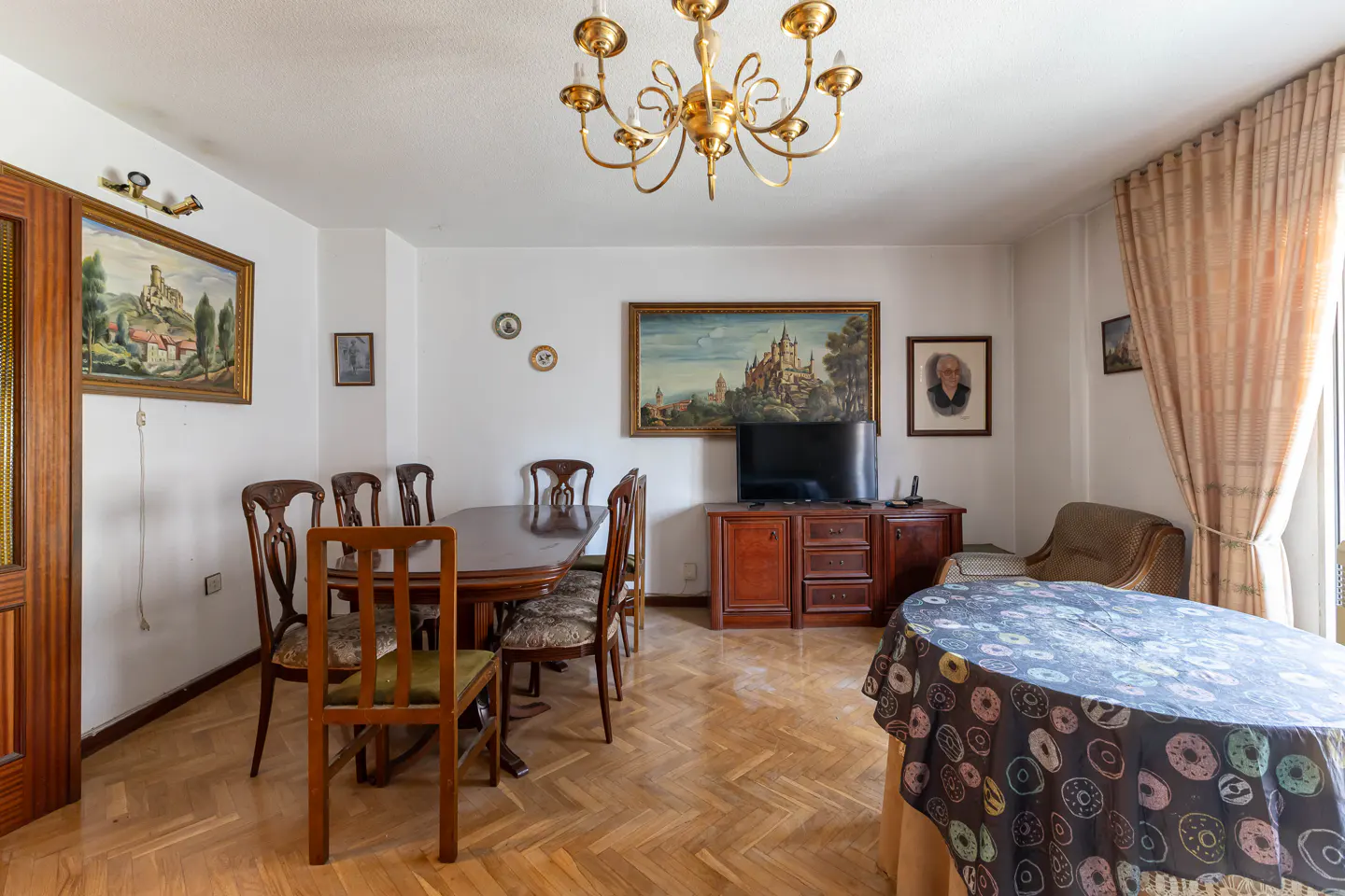 A dining room with a wooden table, chairs, and parquet floor. A TV sits on a wooden cabinet. Paintings adorn the white walls. A gold chandelier hangs from the ceiling.
