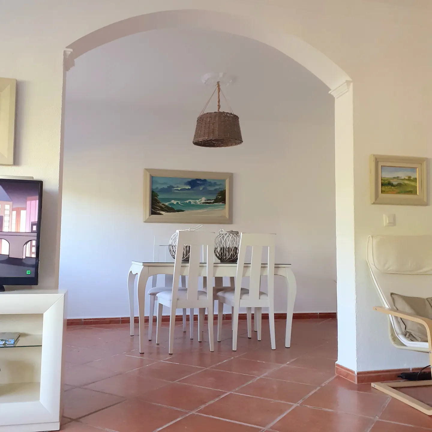 Bright dining room with white table and chairs, terracotta tile floor, and arched doorway. A woven basket light fixture hangs above.