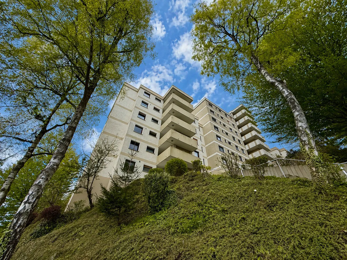 Low-angle shot of a beige apartment building with balconies, framed by green trees against a blue, cloudy sky.
