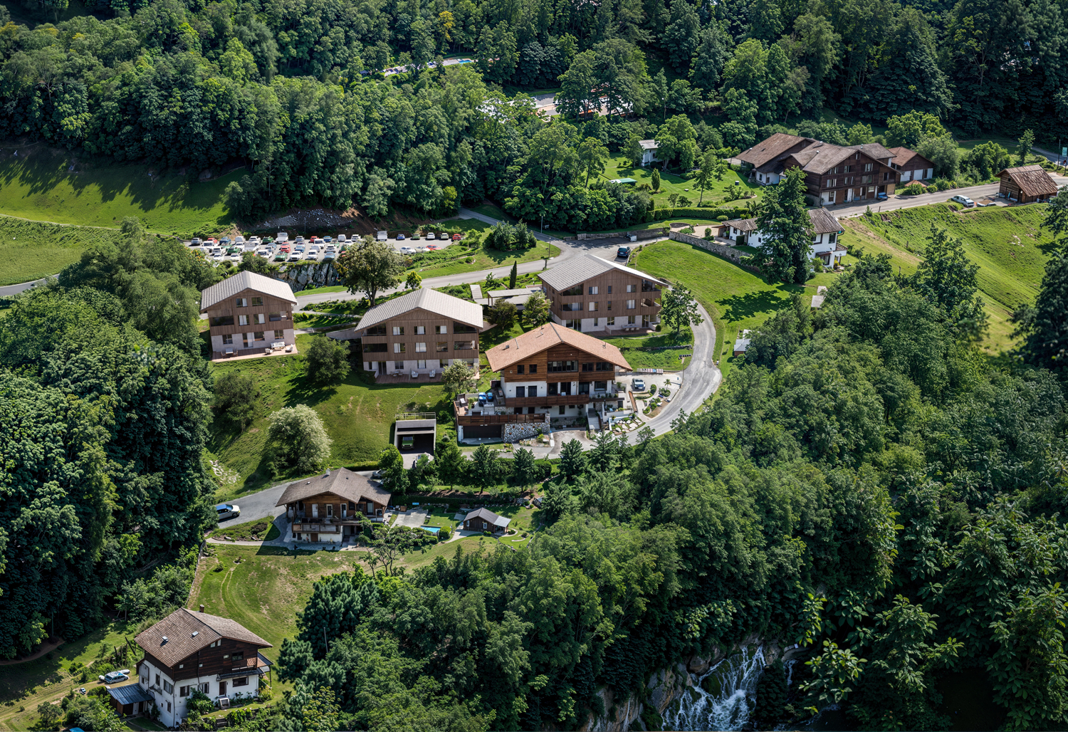 Aerial view of several wooden chalet-style houses nestled in a lush green, forested landscape with a small waterfall. [AI-generated text]