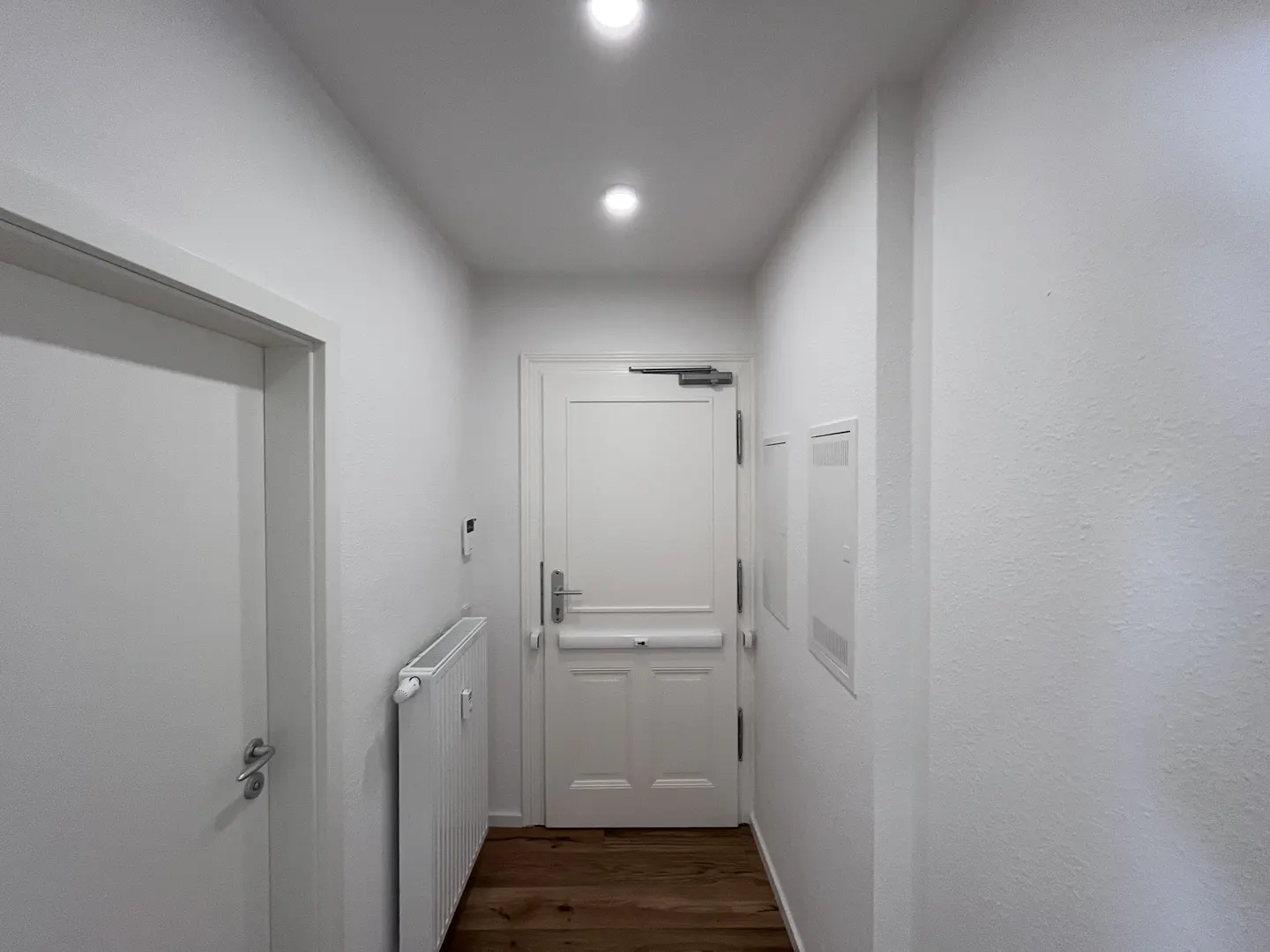 Hallway with white walls, wood floor, and two white doors. One door has a silver handle and a white radiator is next to it.