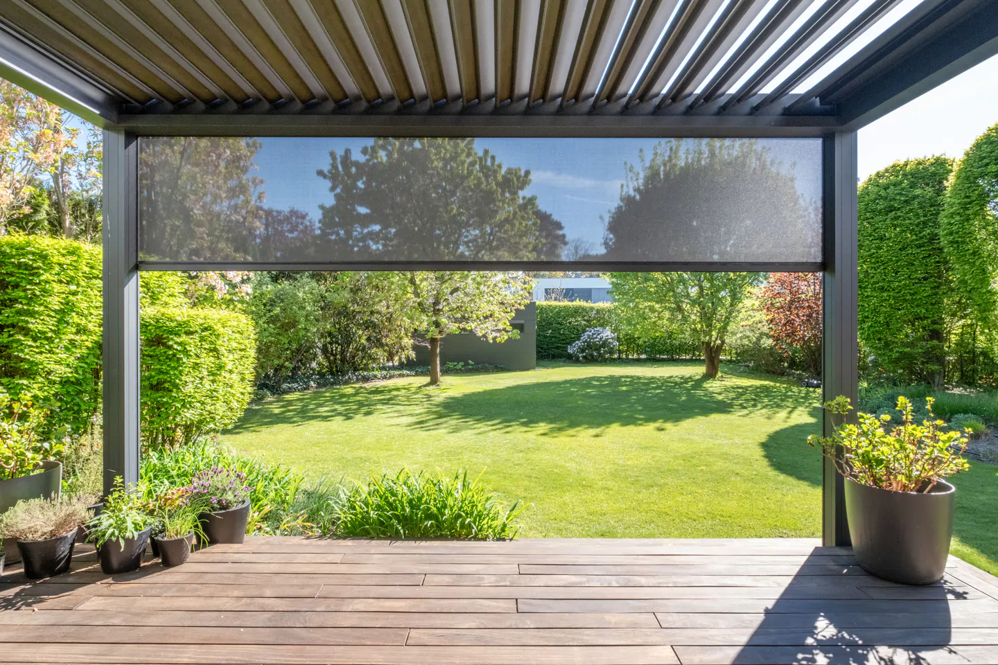 View from a wooden deck under a pergola with a lowered sunshade, overlooking a green lawn and trees.