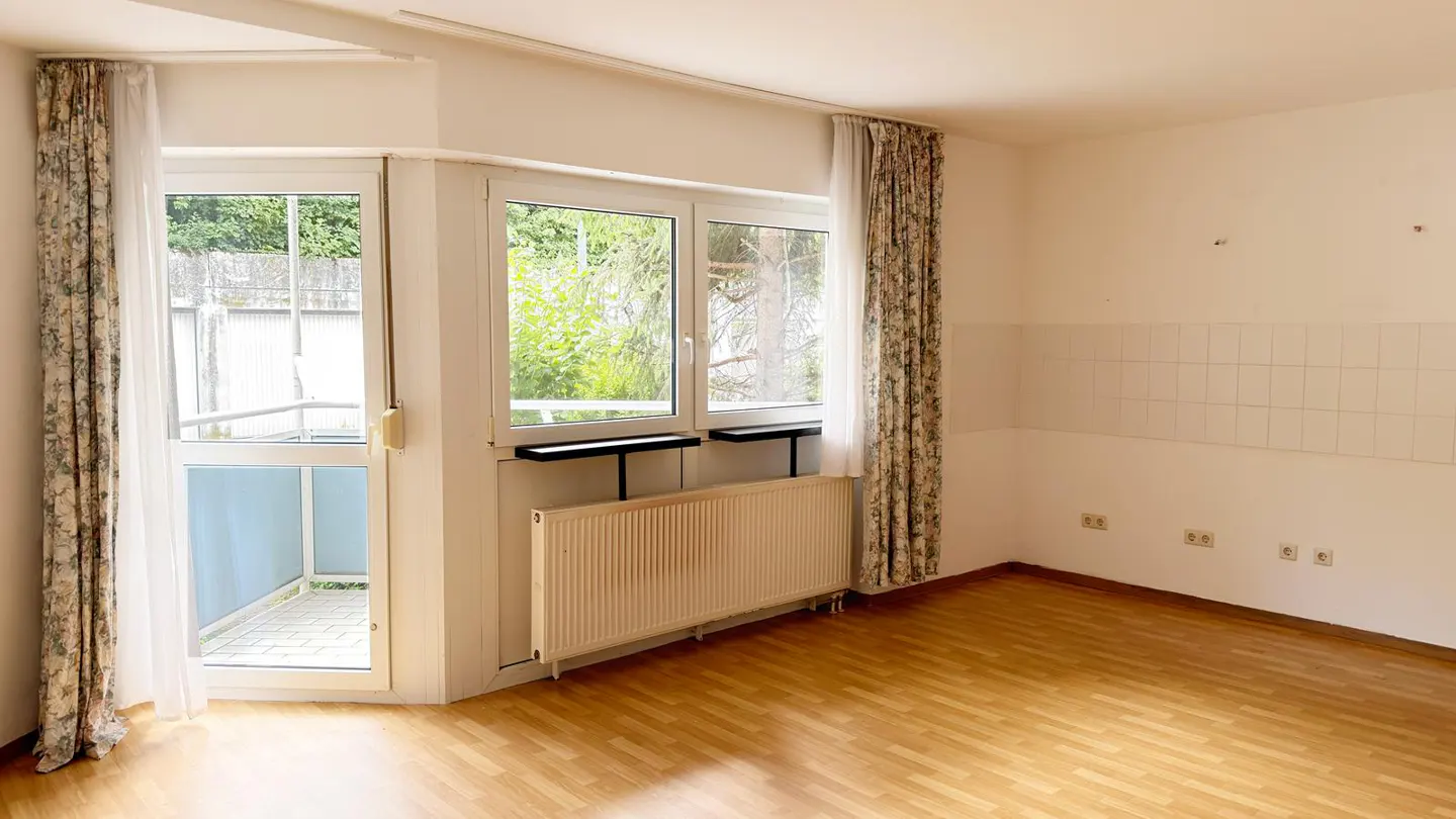Bright, empty room with wood floors, white walls, and floral curtains. Windows overlook greenery, and a door leads to a balcony.