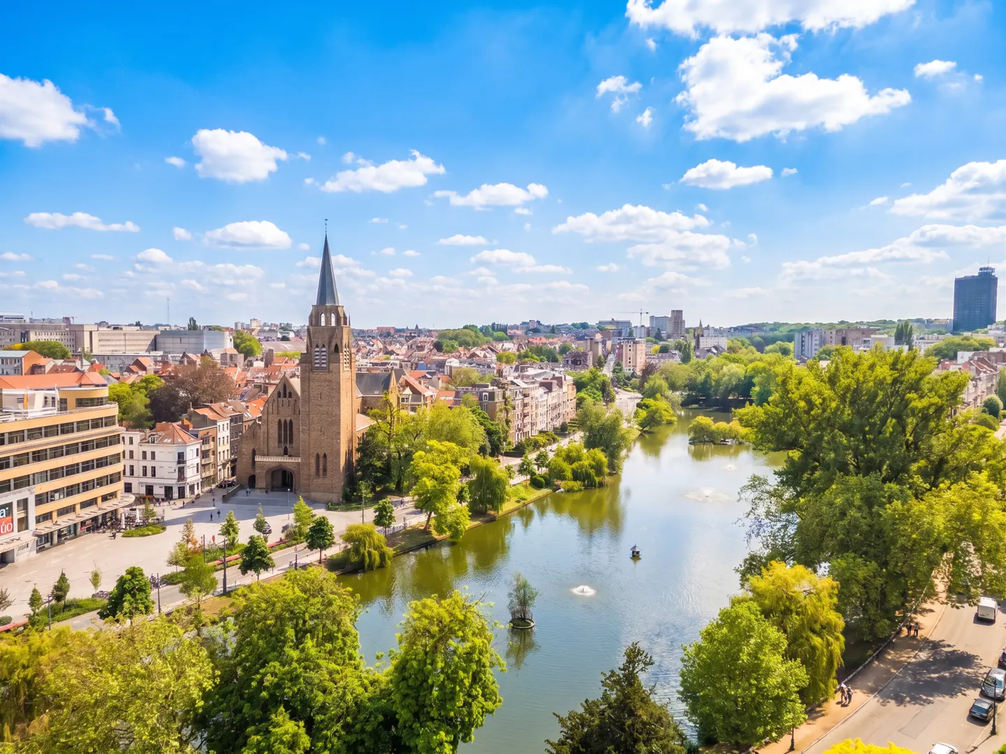 Aerial view of Brussels, Belgium, featuring a lake, church with a tall spire, and buildings under a blue sky with white clouds.