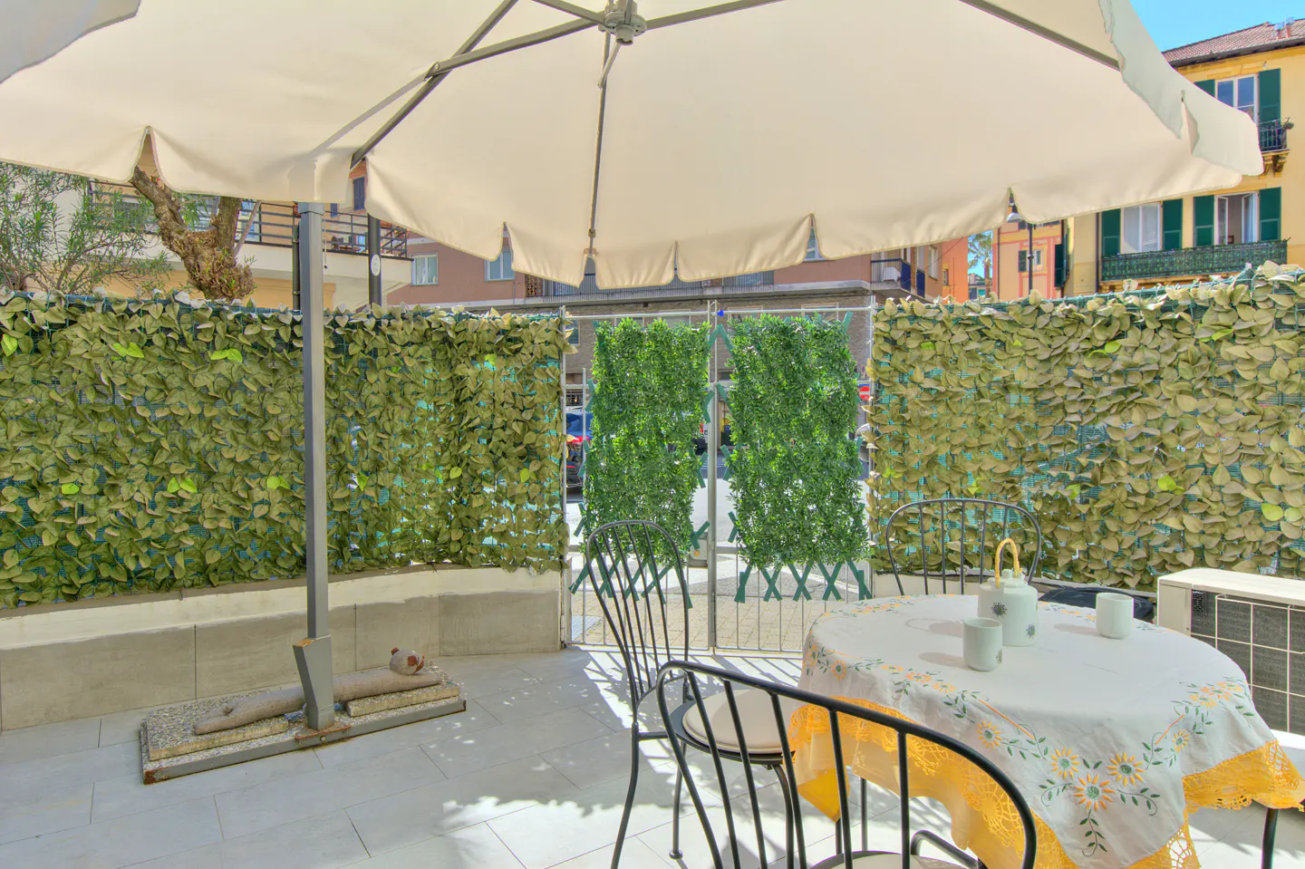 Outdoor patio with a white umbrella, table with a floral tablecloth, and black chairs. Green leafy privacy screens surround the area.