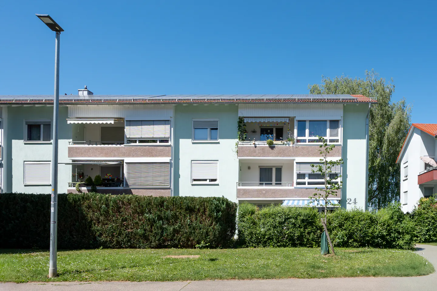 A light green three-story apartment building with balconies, solar panels on the roof, and a hedge in the front yard.