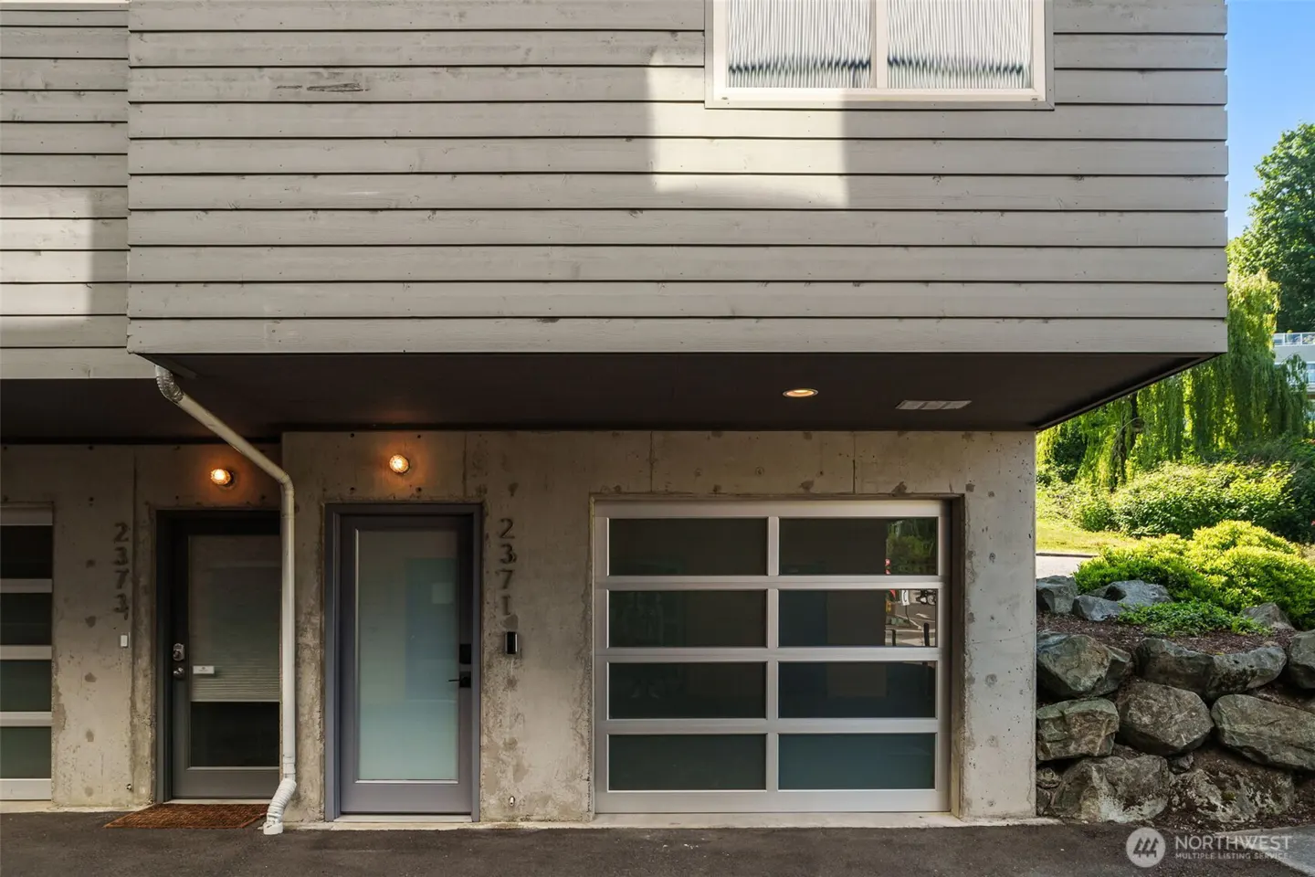 Exterior view of a modern townhouse with gray wood siding, concrete base, glass garage door, and front door. Address number 2371 is visible.