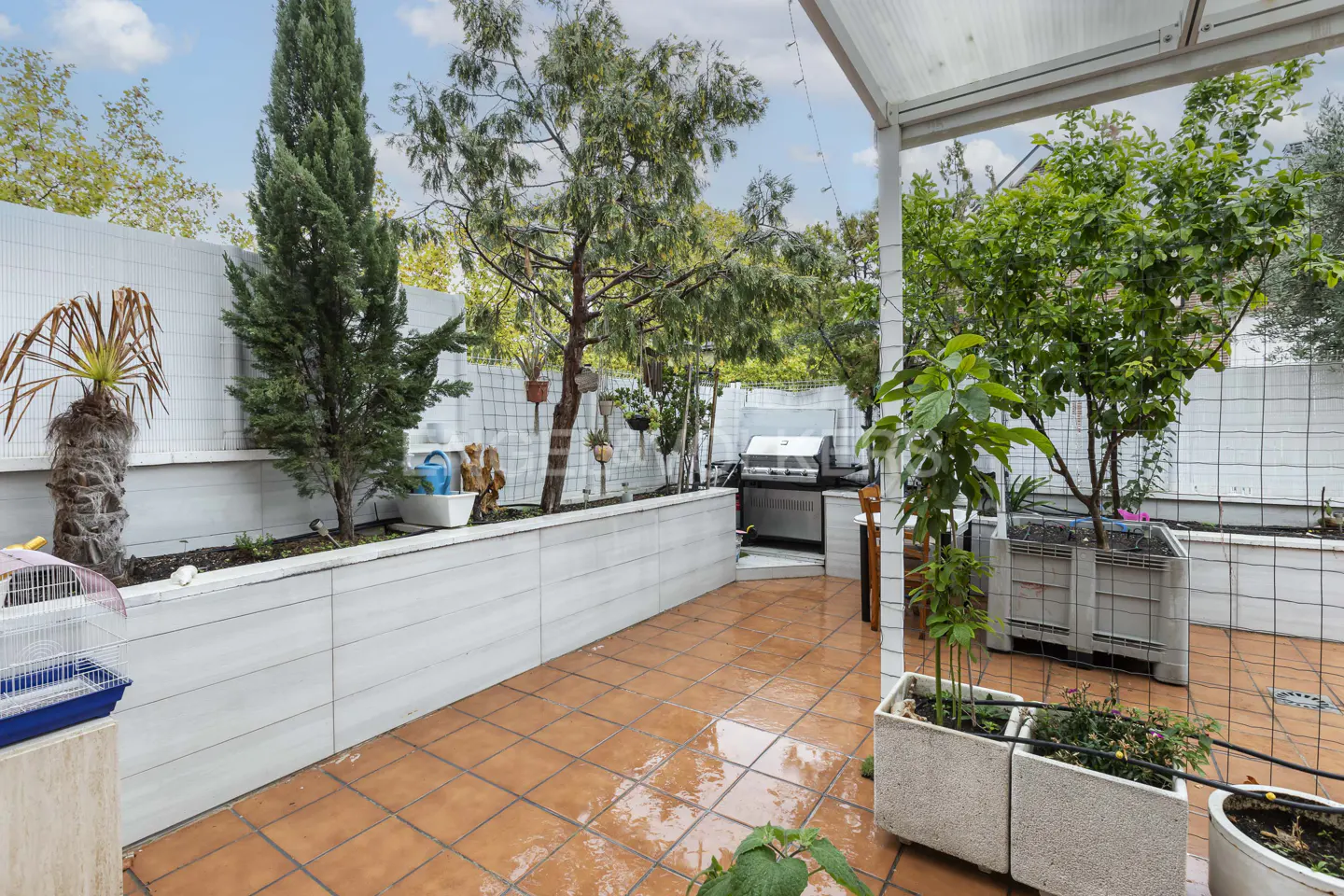 A tiled patio with a white raised garden bed, various potted plants, a small grill, and a birdcage under a partial roof.