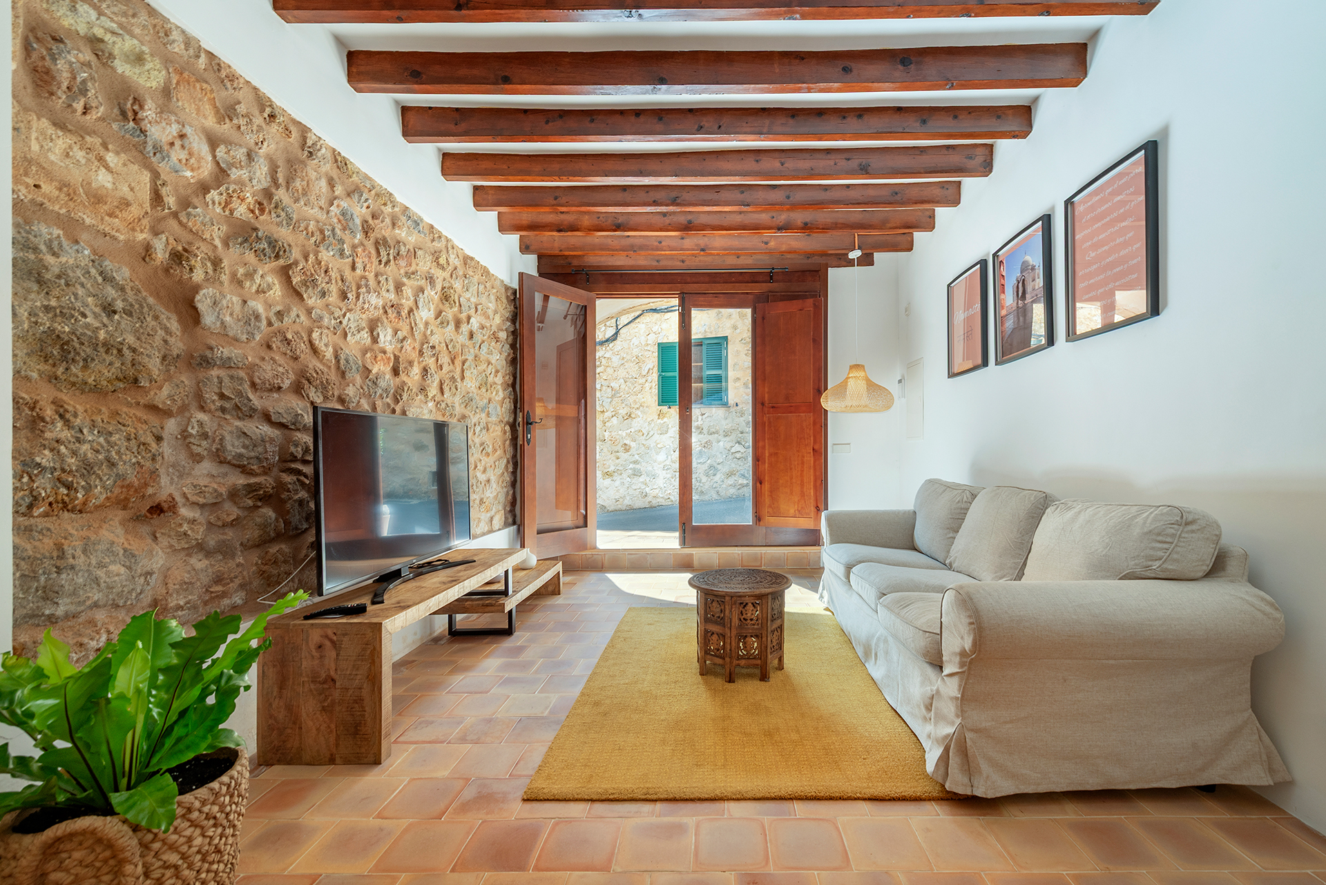 Living room with stone wall, wooden ceiling beams, beige sofa, yellow rug, and open doors leading to a stone courtyard. [AI-generated text]
