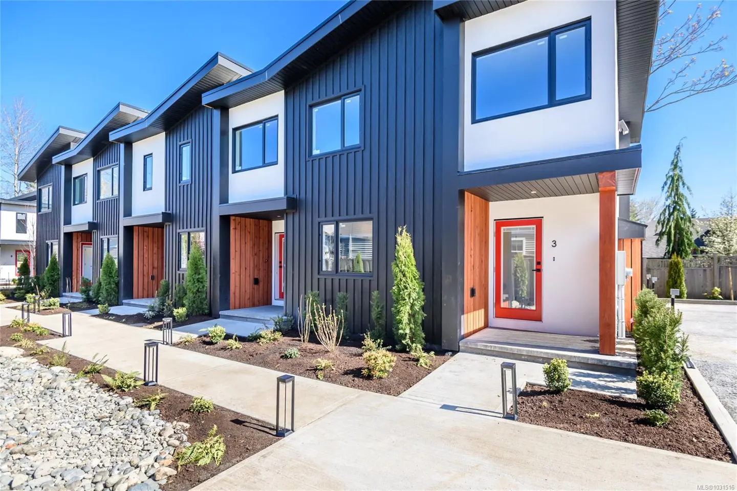 Row of modern townhouses with black siding, white accents, and red doors. A concrete walkway leads to each unit, landscaped with small plants.