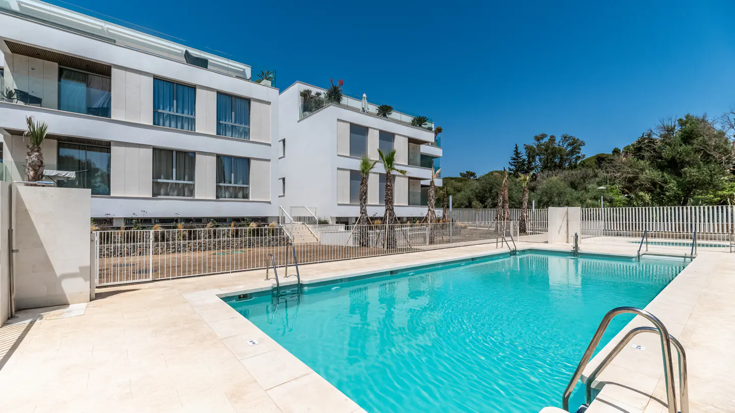 A bright blue swimming pool sits in front of modern white apartments under a clear blue sky.