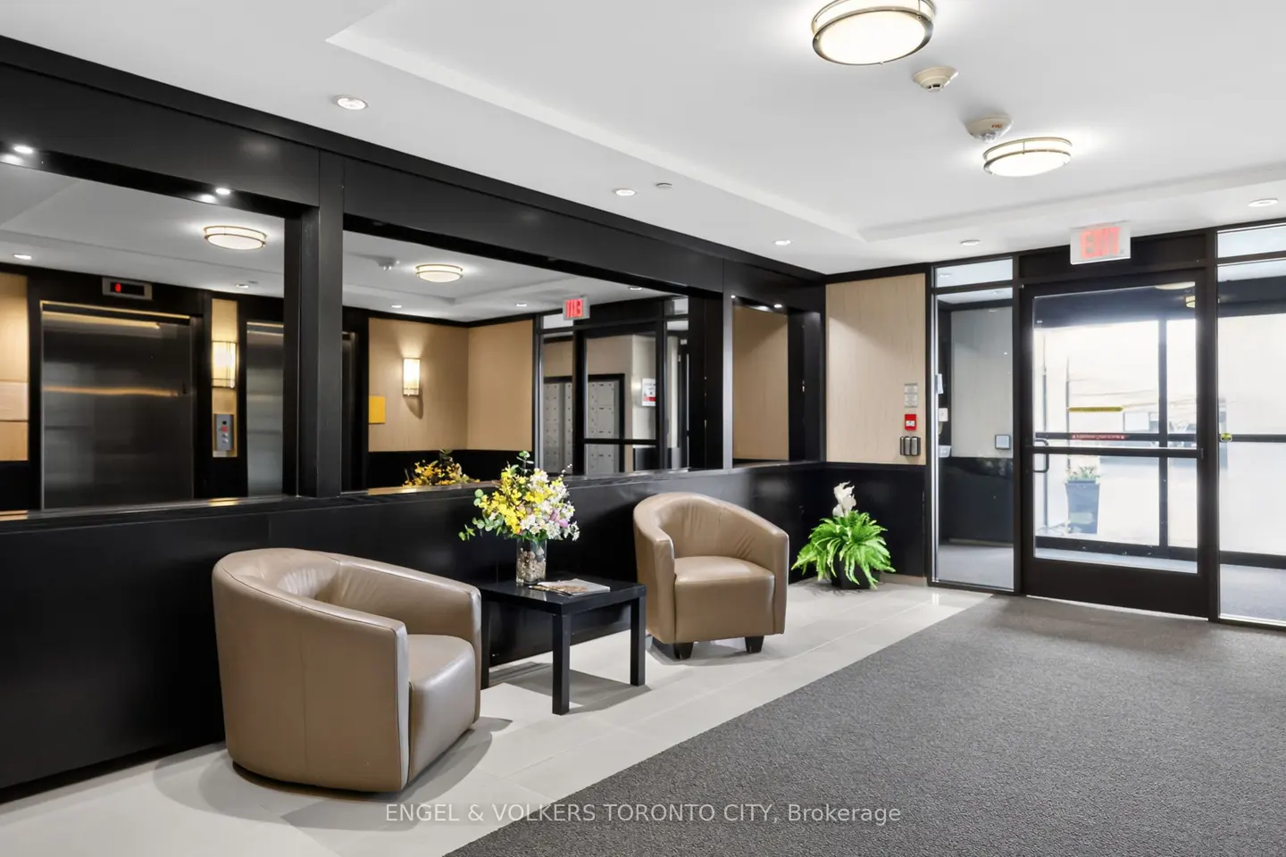 Modern building lobby with two beige armchairs, a small black side table with flowers, and glass doors leading outside.
