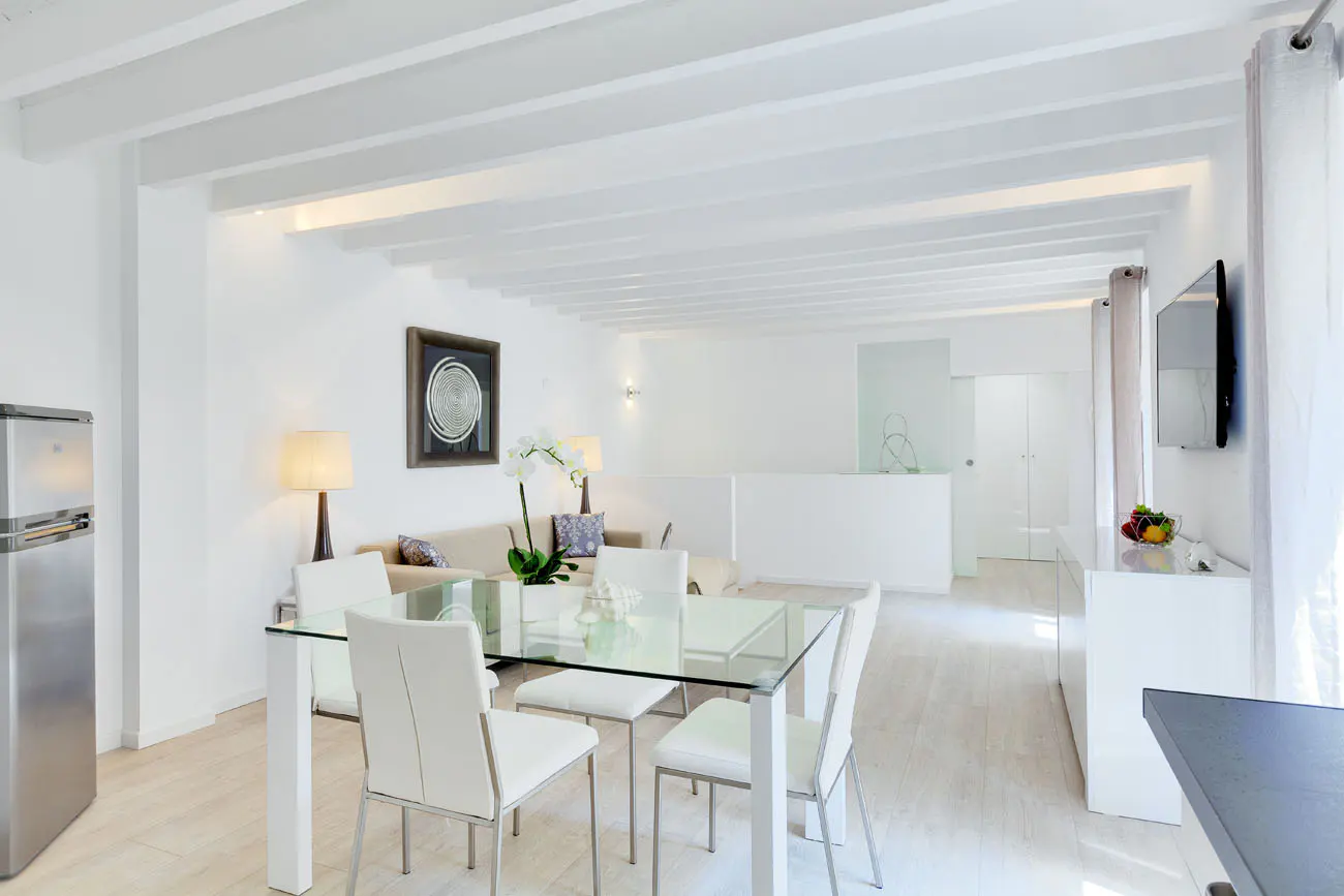 Bright, open-concept living space with white walls, beamed ceiling, and light wood floors. A glass table with white chairs sits near a stainless steel refrigerator.