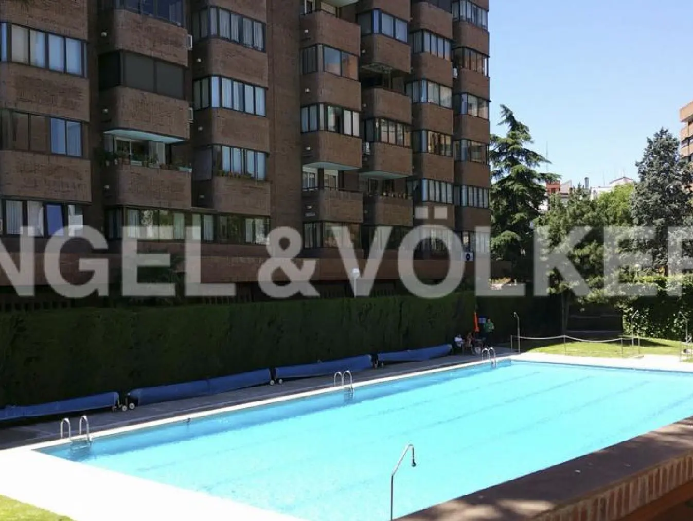 Outdoor pool with blue water in front of a brown brick apartment building and green hedges.